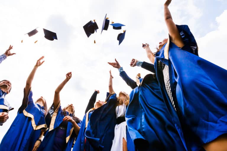 happy young graduates in graduation gowns throwing graduation caps into the air