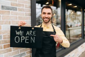 how to start a business with no money open sign Welcome, we are open. Portrait of caucasian attractive male waiter standing outdoors near a restaurant or cafe, points a finger at a signboard OPEN and friendly smile. Support small business concept