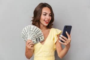 get paid to text Portrait of a smiling young girl in dress standing over gray background, holding mobile phone, showing money banknotes