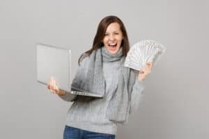 How To Make 300 Dollars Fast Overjoyed woman in sweater working on laptop pc computer hold lots bunch of dollars banknotes cash money isolated on grey background. Healthy lifestyle online treatment consulting cold season concept