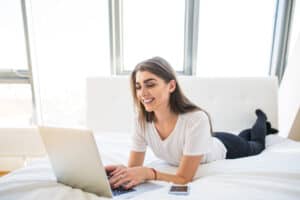 jobs for lazy people Portrait of beautiful happy young woman lying in bed and using laptop at home