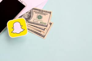 make money on Snapchat NY, USA - DECEMBER 26, 2019: Snapchat paper logo lies with envelope full of dollar bills and smartphone
