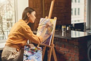 hobbies that make money, Charming young woman sitting by the window and drawing abstract eye with paints in home art studio