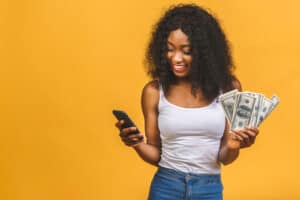 Is Blackout Bingo Legit Happy winner. Portrait of african american successful woman 20s with afro hairstyle holding lots of money dollar banknotes and using phone isolated over yellow background.