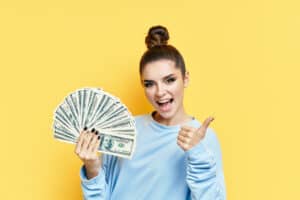 Side Hustles That Pay Weekly Happy excited young woman showing money banknotes with thumbs up on yellow background. Motivation, success concept