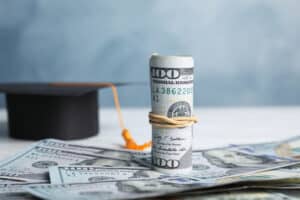 Easy Degrees That Pay Well Dollar banknotes and student graduation hat on white wooden table against blue background. Tuition fees concept